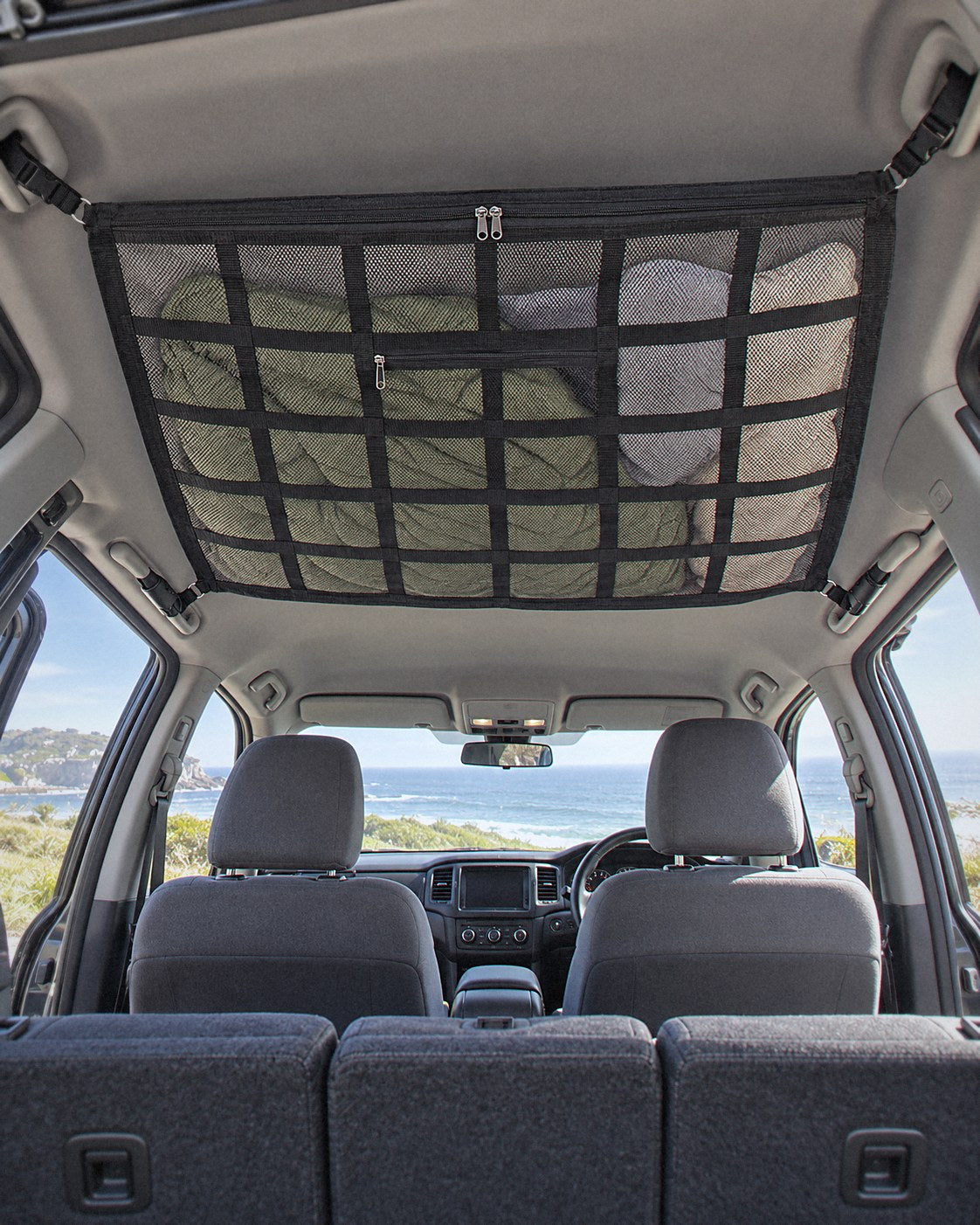 Stone cargo net loaded with a jacket and pillow, strapped to the roof grab handles of an SUV, rear-view mirror visible below
